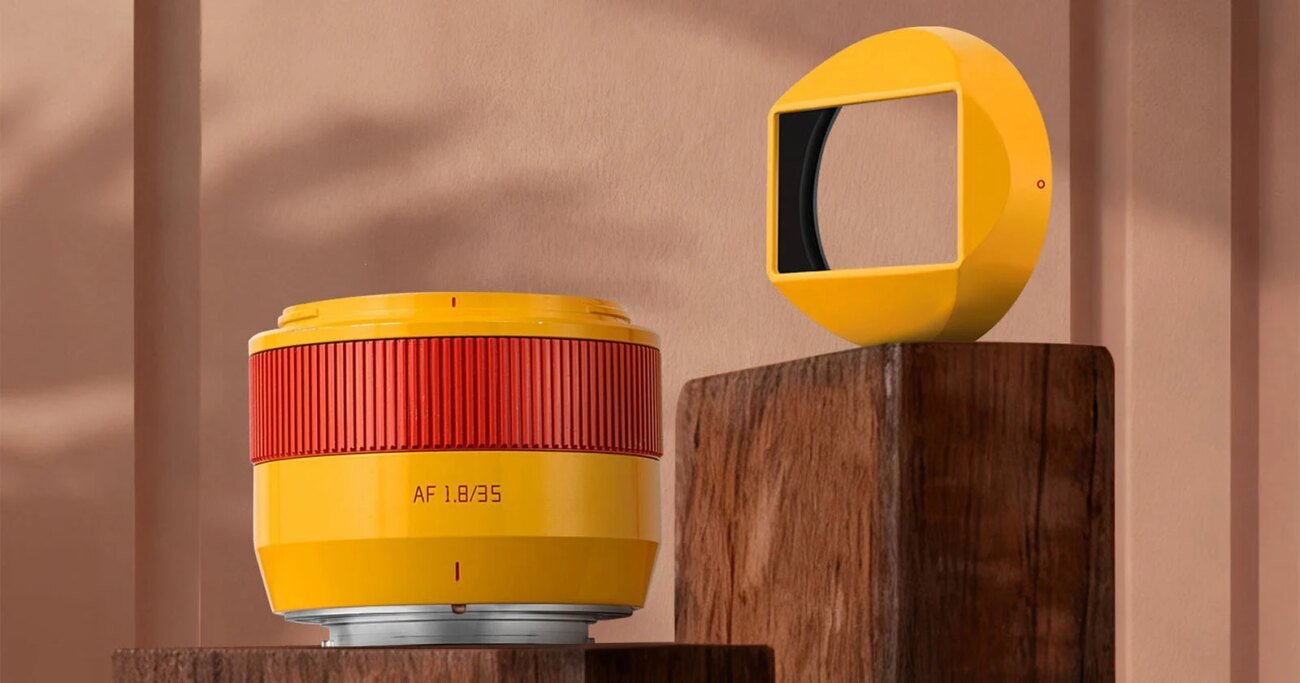 A yellow camera lens with a red ring and "AF 1.8/85" text sits on a wooden block next to a matching yellow geometric object with a rectangular black opening, against a beige background.